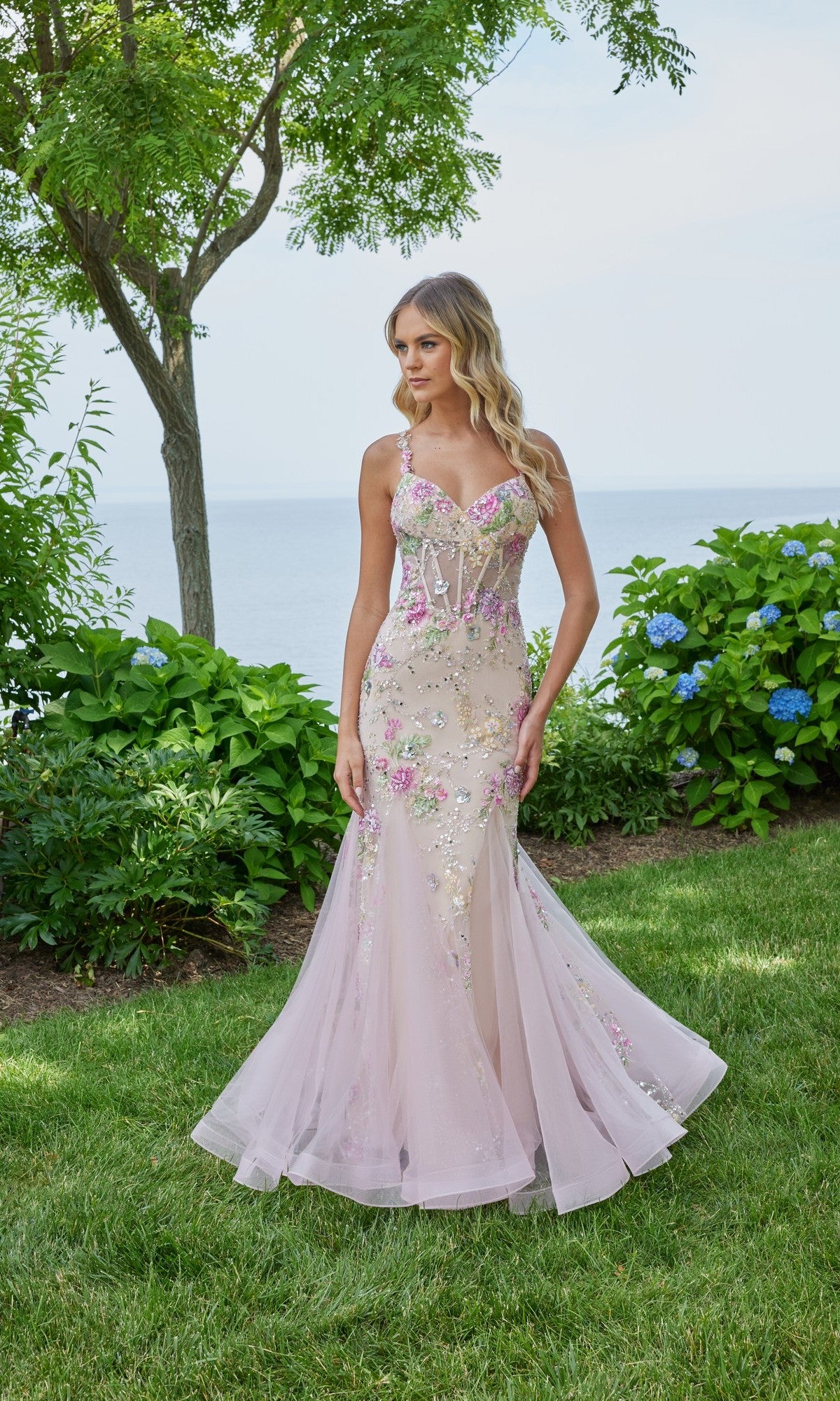 A woman stands on grass in front of bushes, wearing the Morilee 49130 long prom dress in blush/multi with a fitted bodice and sheer, floral-embellished skirt. Trees and water appear in the background.